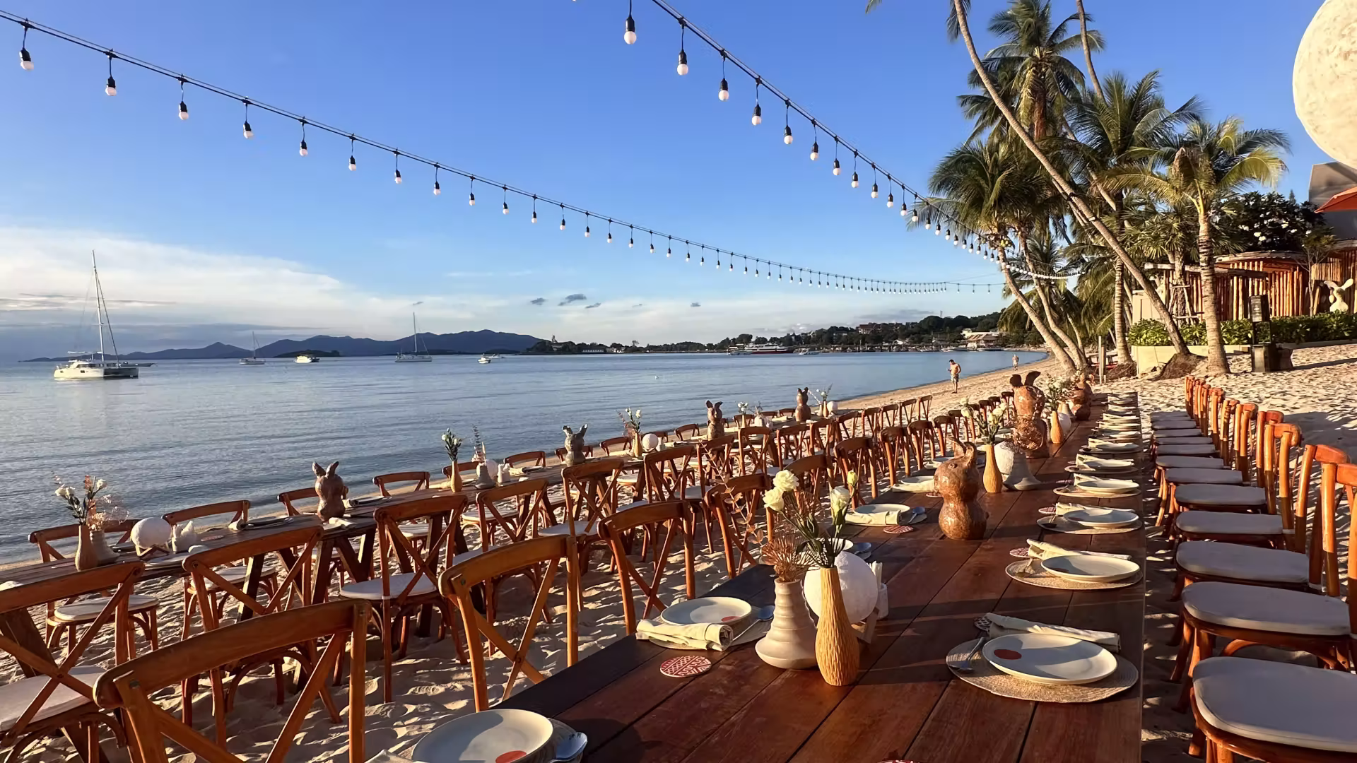 Long wooden tables with place settings set up on a sandy beach under string lights, palm trees, and clear blue sky near calm sea with anchored boats.
