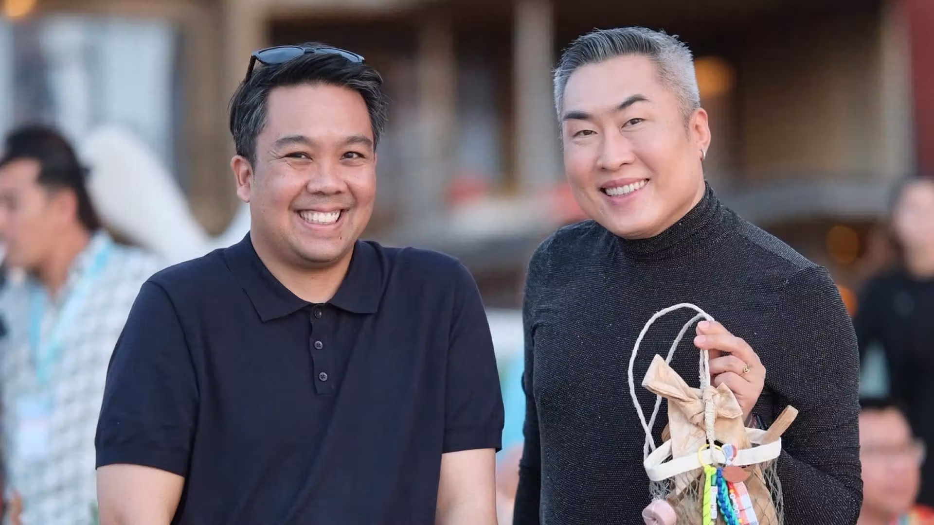 Two smiling men posing at an outdoor event, one wearing a navy polo and the other in a dark long-sleeve top with a yellow skirt holding a decorative basket.