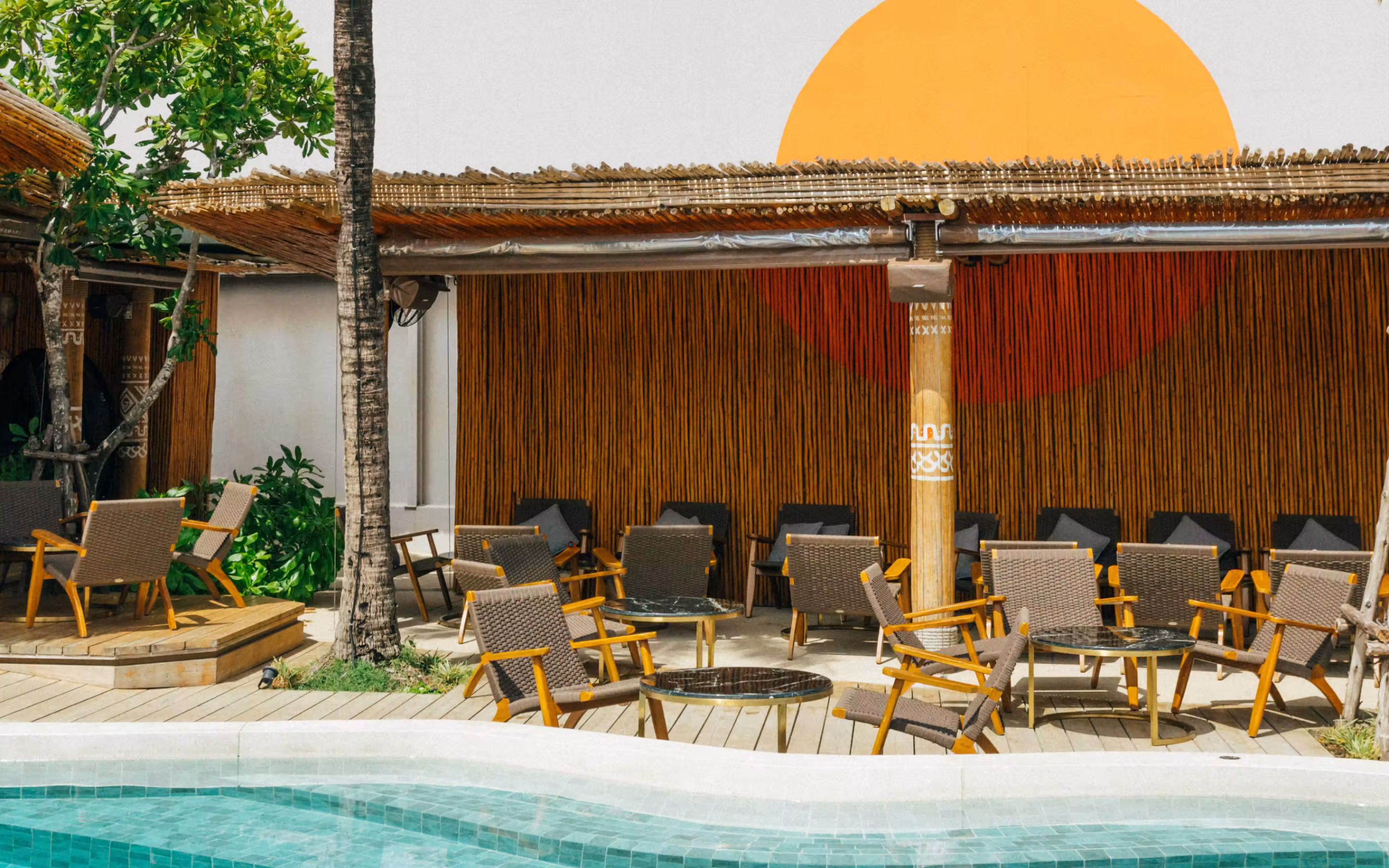 Outdoor poolside seating area with woven chairs and round tables under a bamboo canopy backed by a wooden fence with a sun mural.