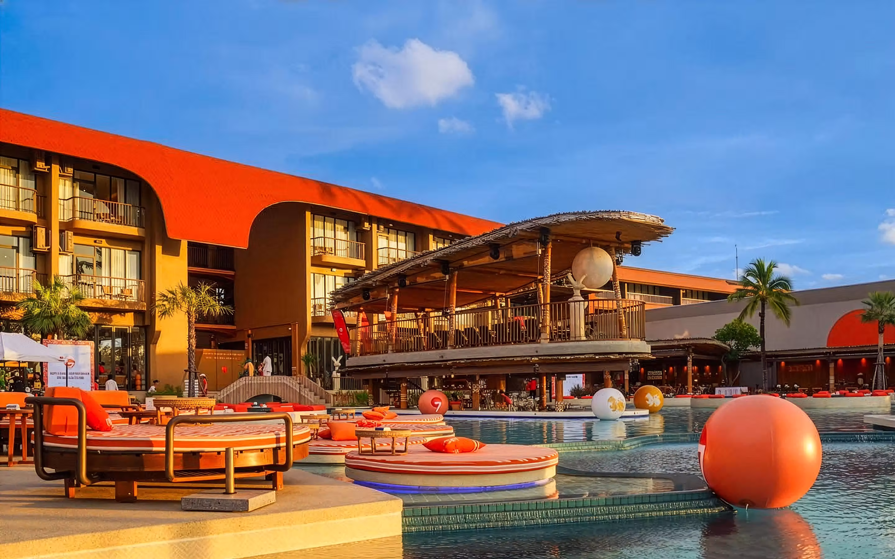 Modern resort pool area with round orange loungers, large decorative balls, and a thatched roof bar under a clear blue sky.