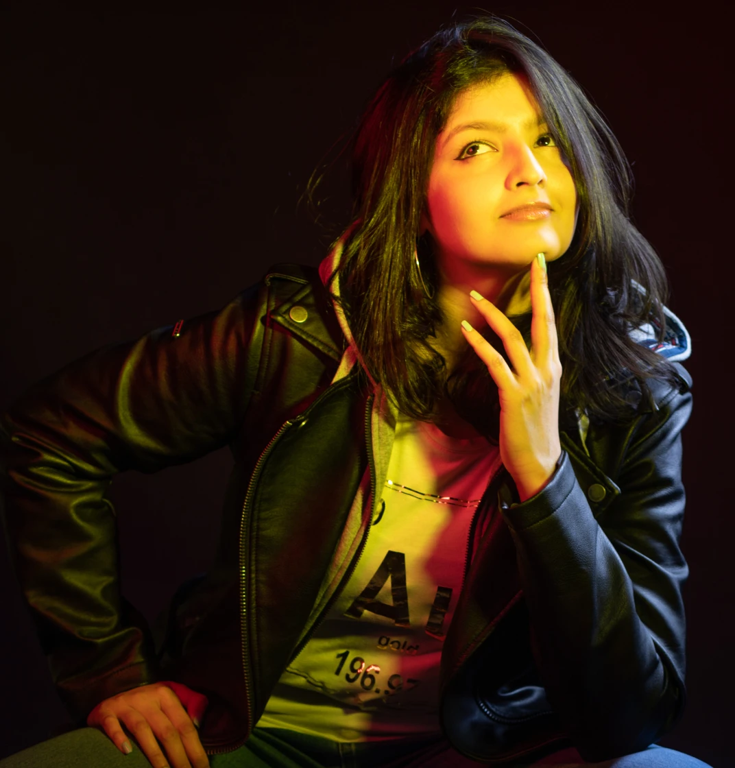 Samia Bukhari photo in black leather jacket illuminated with yellow and red lighting, posing thoughtfully against a dark background.