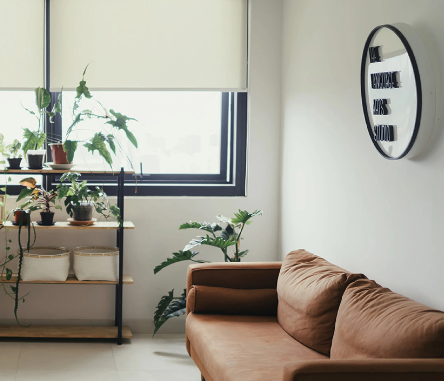 Minimalist studio with a brown sofa, potted plants on shelves and floor, and a round wall sign reading 'The Language Arts Studio'.