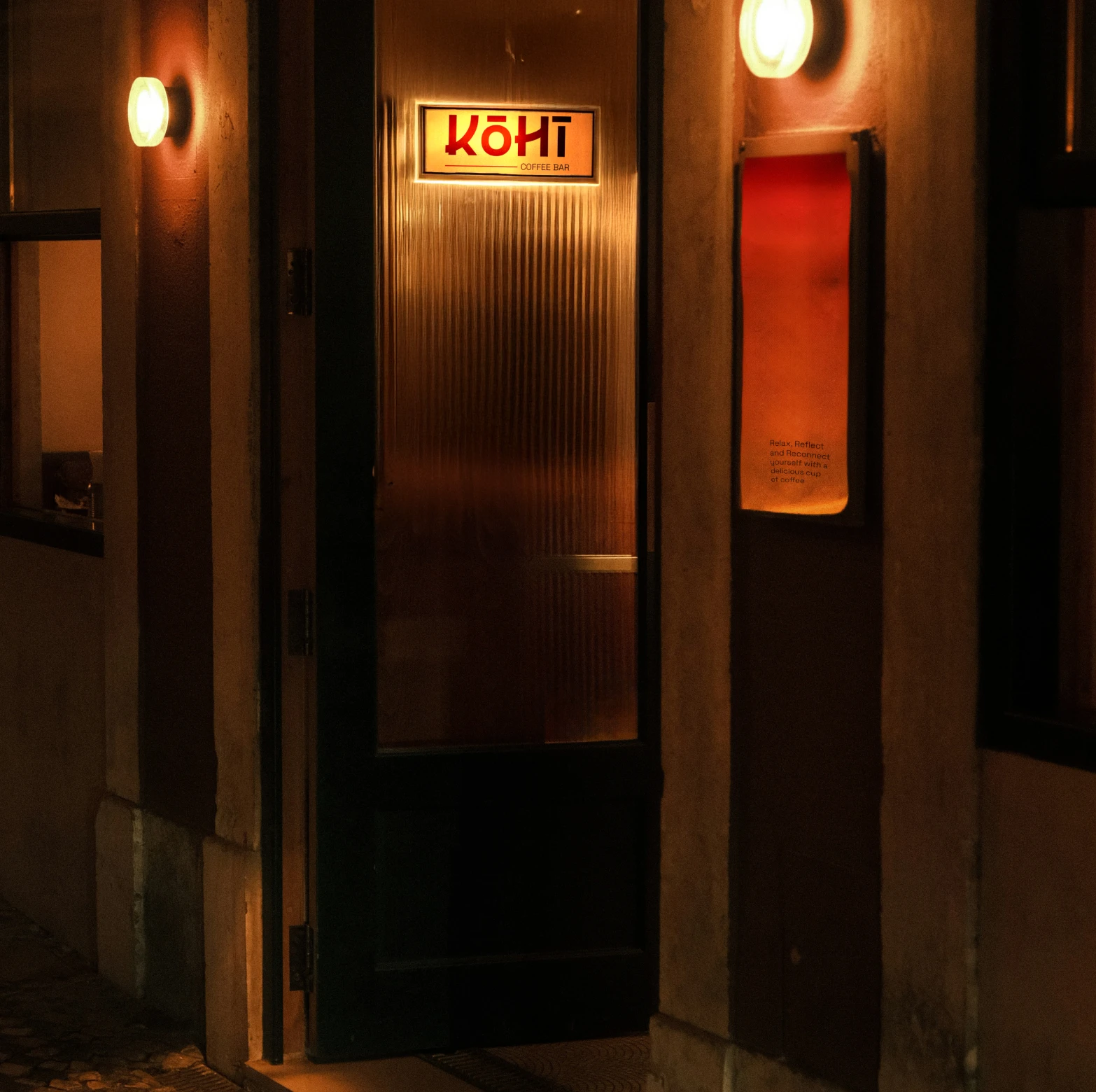 Entrance door of Kōhī Coffee Bar lit by warm wall lights at night.