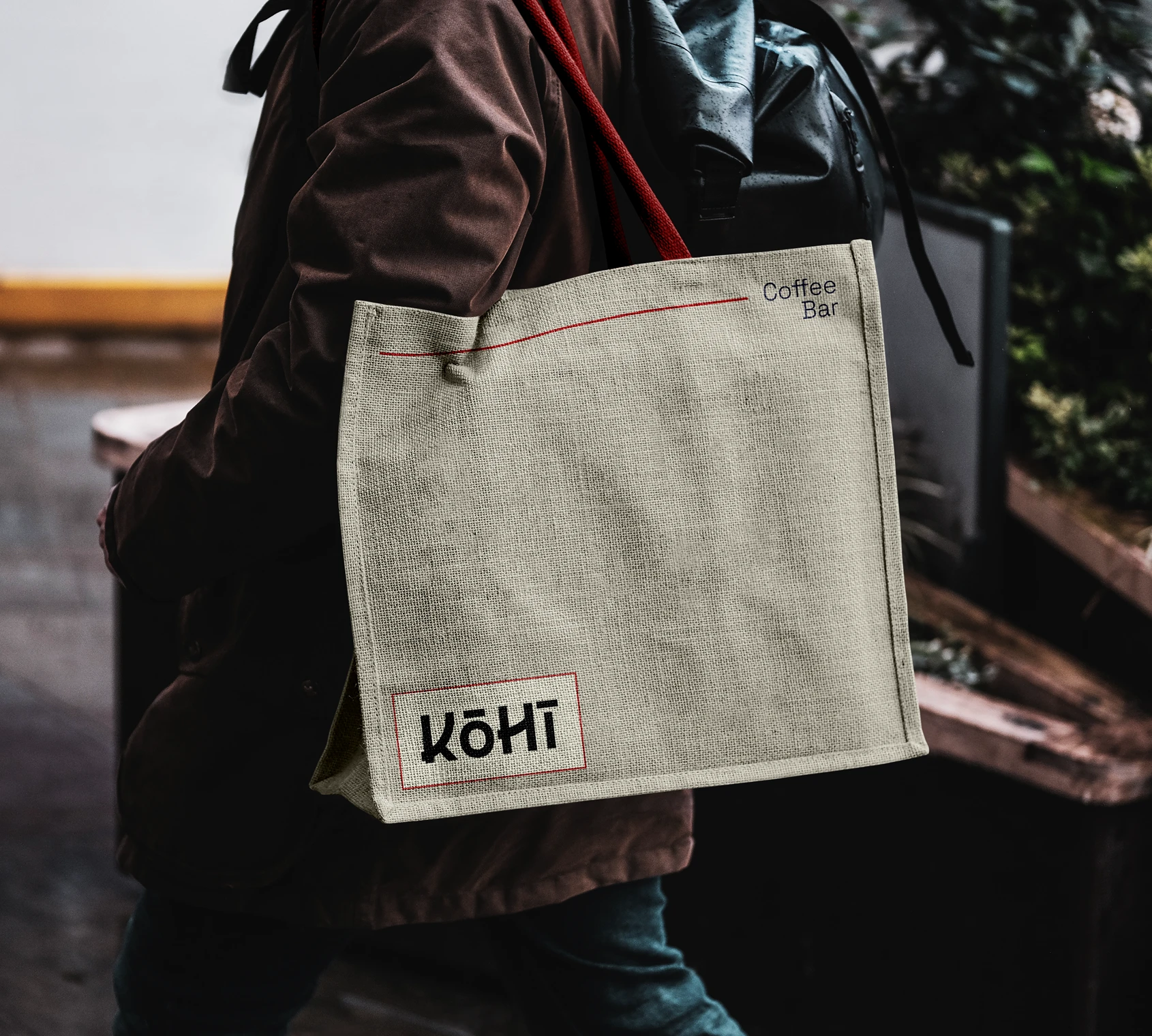 Person wearing a brown jacket carrying a beige tote bag with red handles that says 'Kōhī' and 'Coffee Bar'.