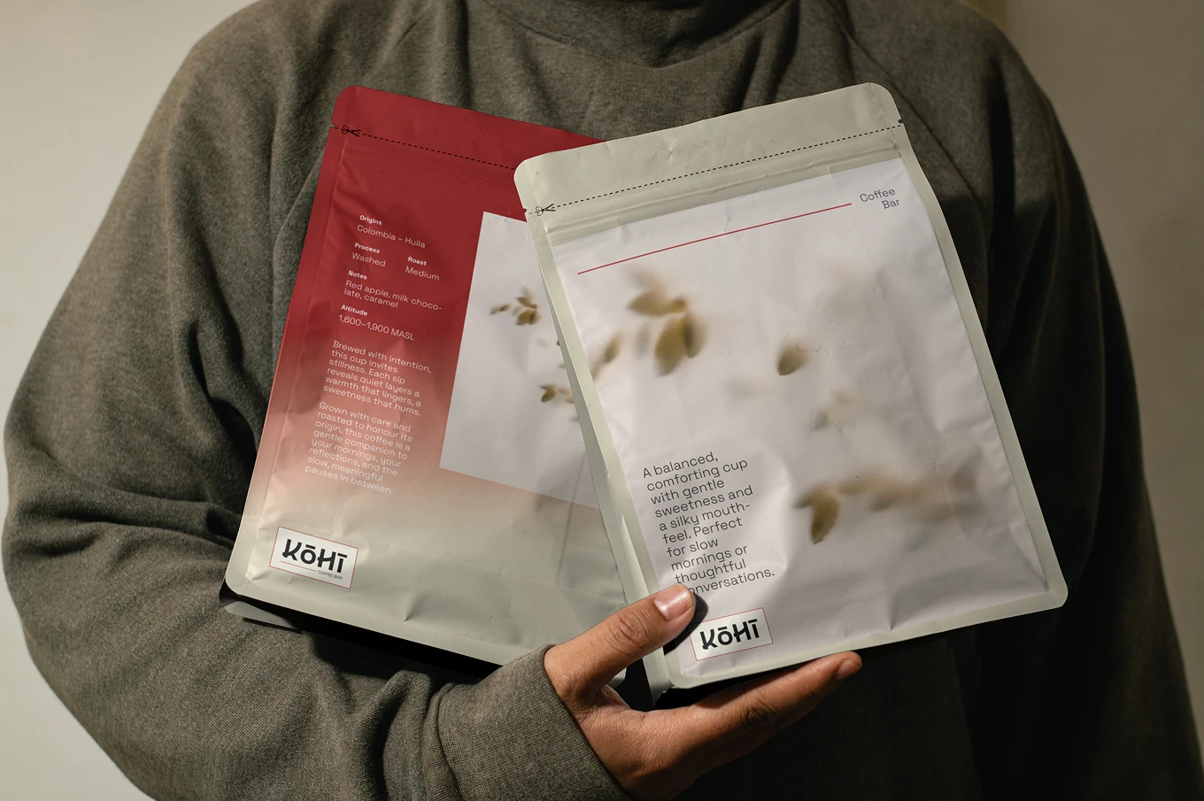 Person wearing a grey sweater holding two packets of Kōhī Coffee Bar coffee, one red and one white with printed descriptions.