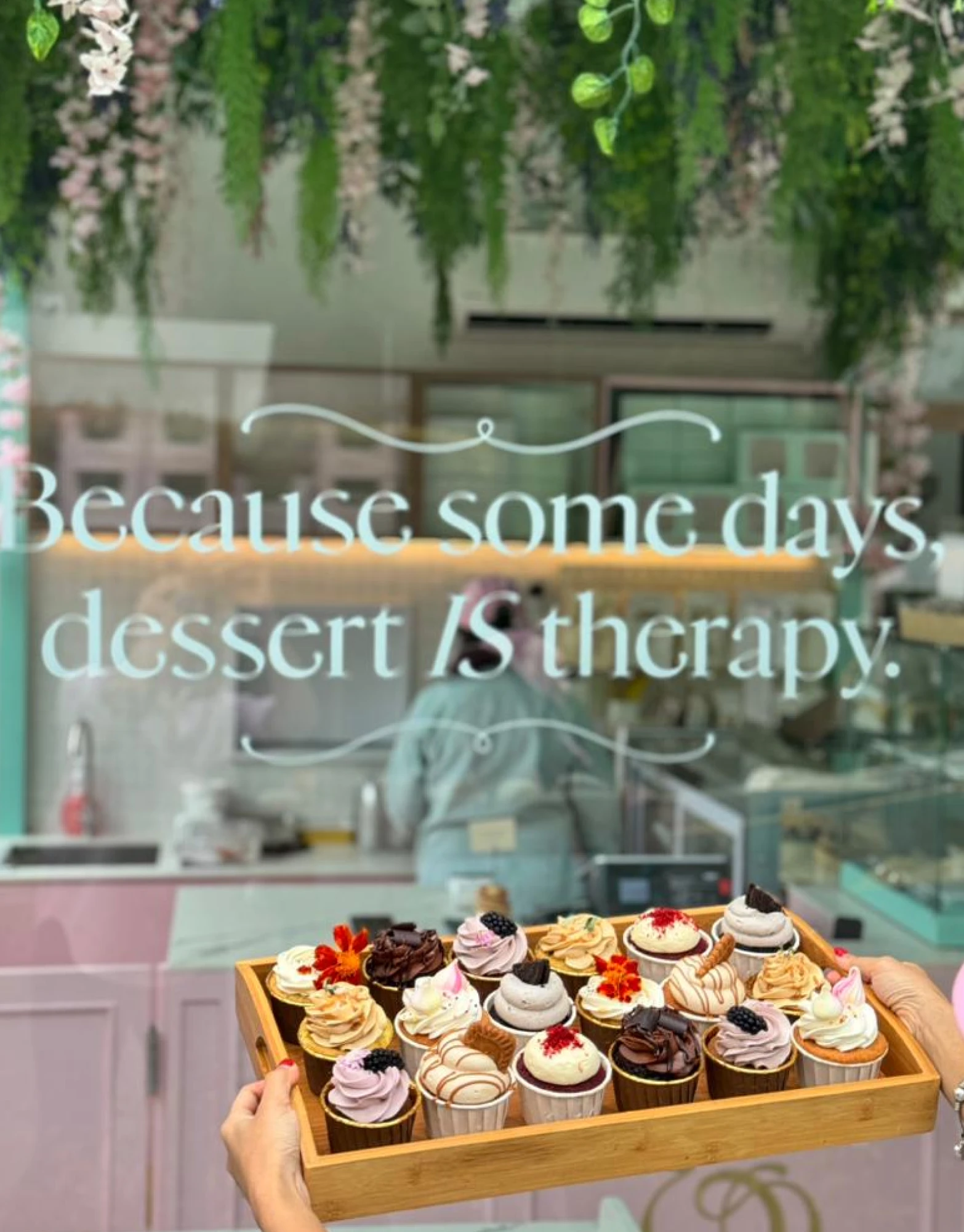 Tray of assorted decorated cupcakes held in front of a window with the phrase 'Because some days, dessert IS therapy' written on it.