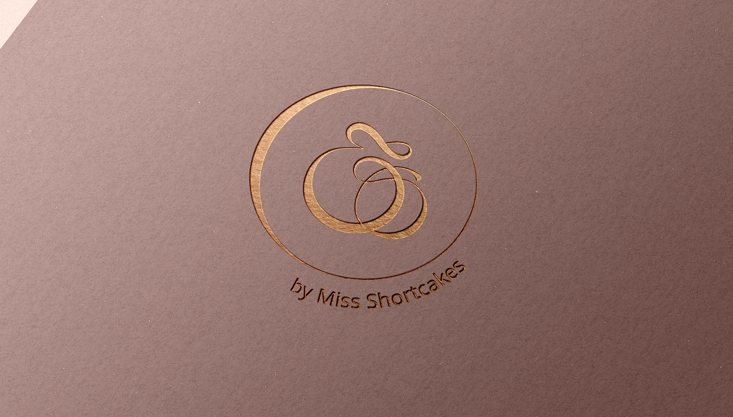 Embossed gold logo with elegant decorative initials and text 'by Miss Shortcakes' on brown paper.