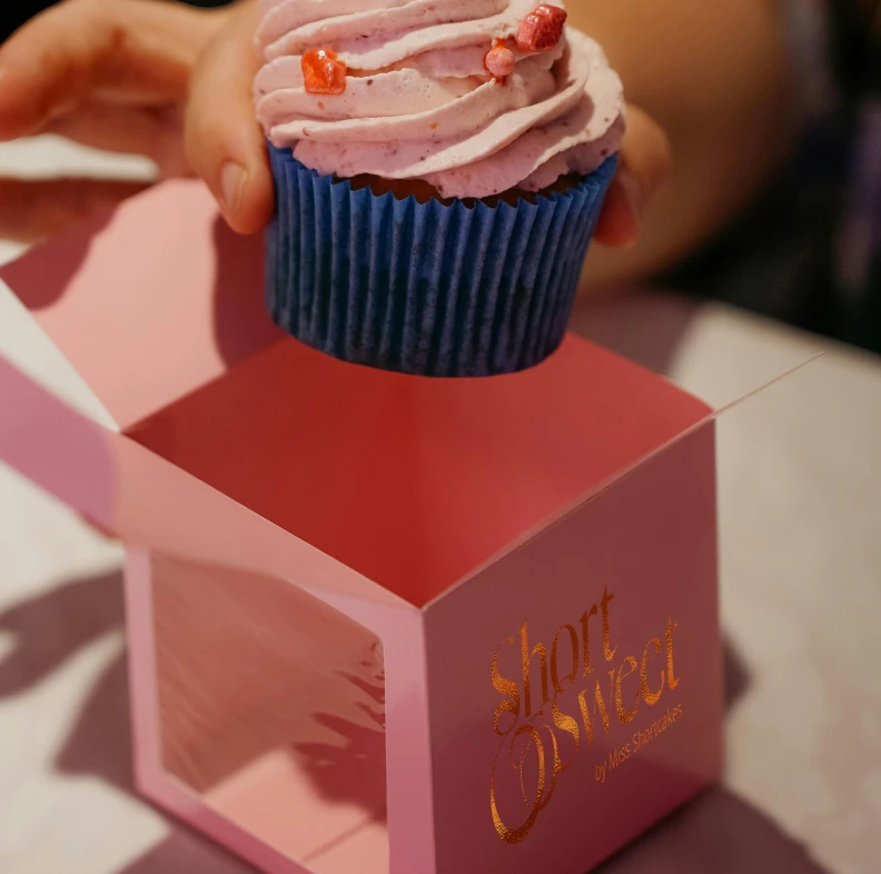 Hand holding a cupcake with pink frosting and red sprinkles above a pink cupcake box labeled 'Short Sweet'.
