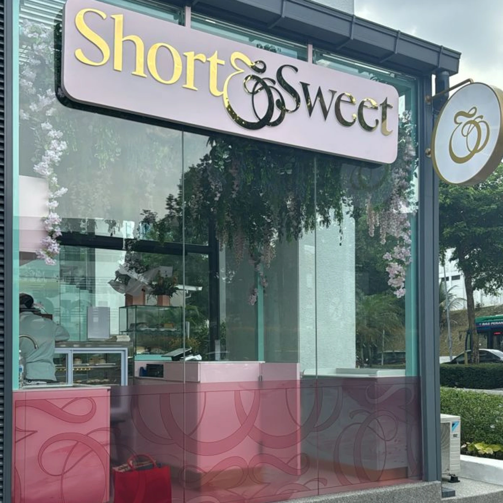 Exterior of a bakery café with a sign reading 'Short & Sweet' and pink counters inside decorated with hanging flowers.