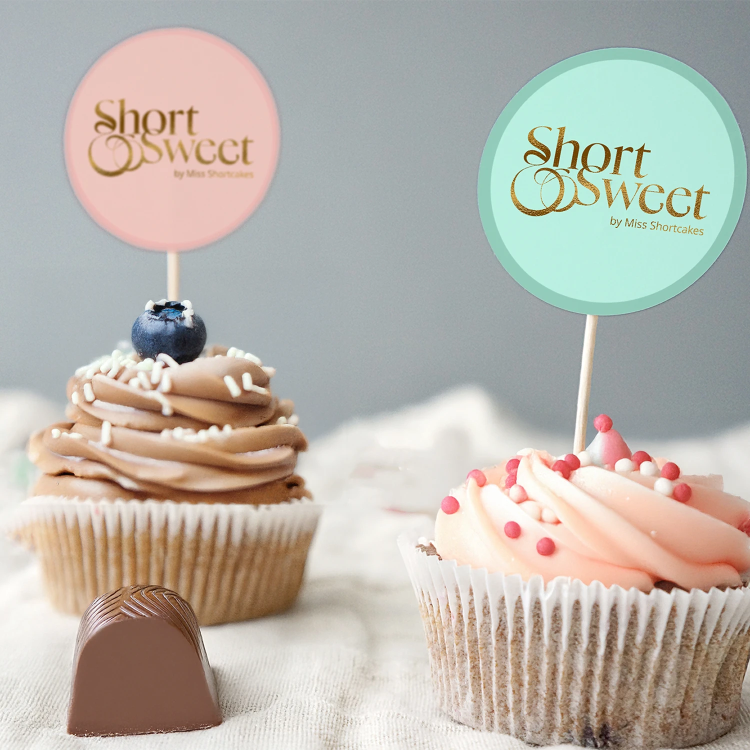 Two decorated cupcakes with swirl frosting and branded toppers reading 'Short & Sweet by Miss Shortcakes' and a piece of chocolate on a cloth surface.
