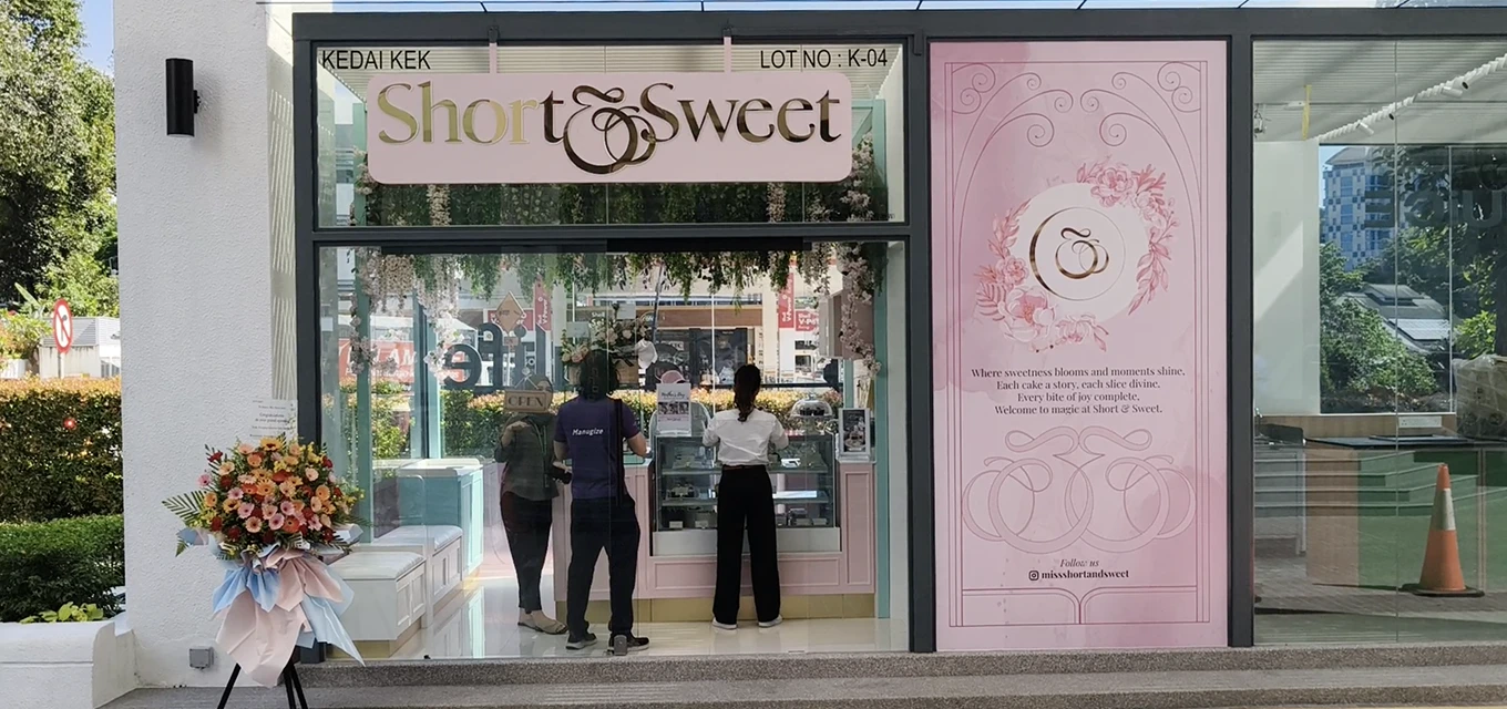 People standing inside a pastel-themed bakery named Short & Sweet, with floral decorations and a pink signboard displaying the bakery's poetic motto.