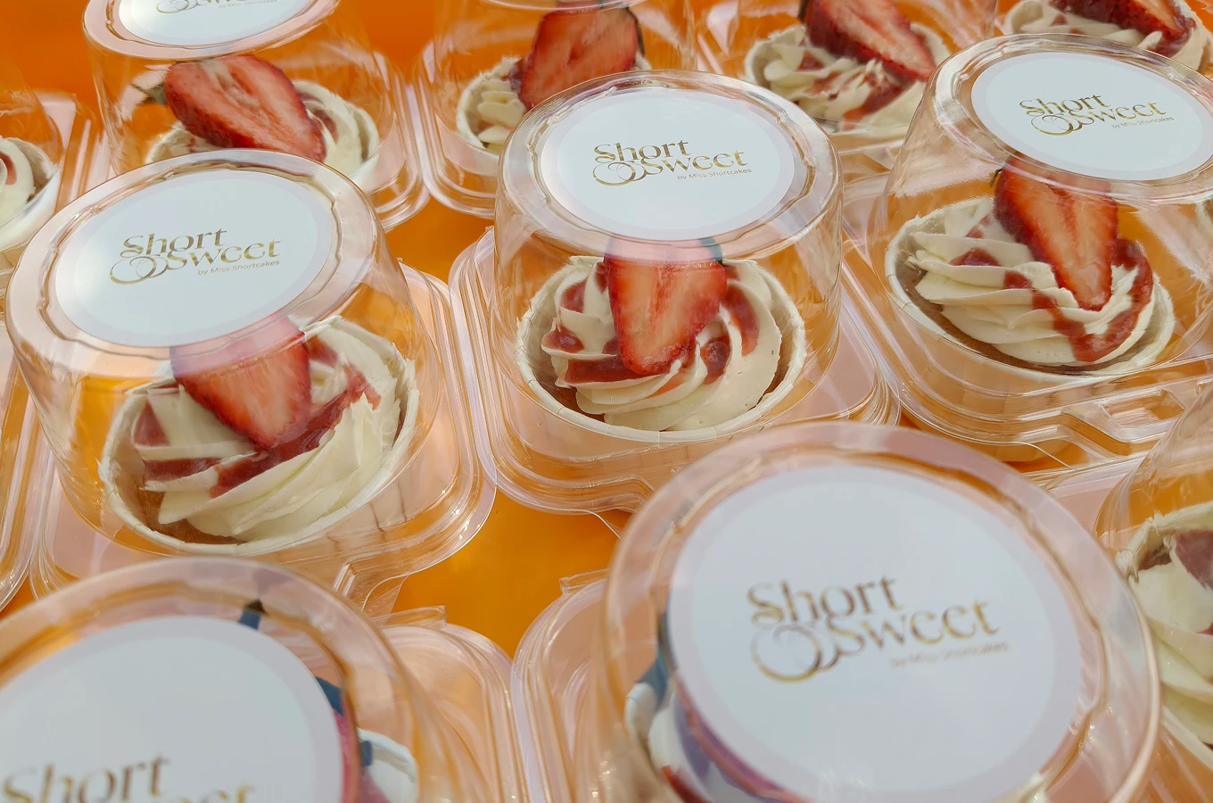 Individual desserts topped with whipped cream, strawberry sauce, and a sliced strawberry in clear plastic containers labeled Short & Sweet.