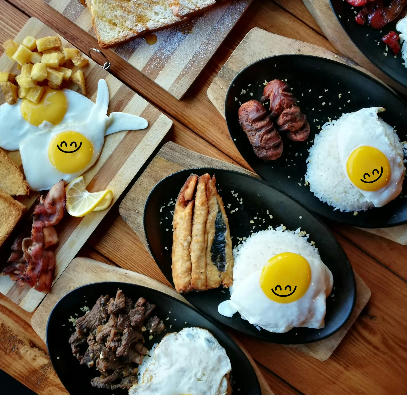 Breakfast spread with fried eggs topped with smiley faces, grilled meats, rice, toast, bacon, potatoes, and lemon wedges arranged on wooden boards and black plates.