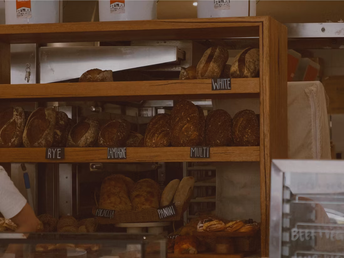 Wooden shelves displaying various types of bread labeled as white, rye, focaccia, and panini in a bakery.
