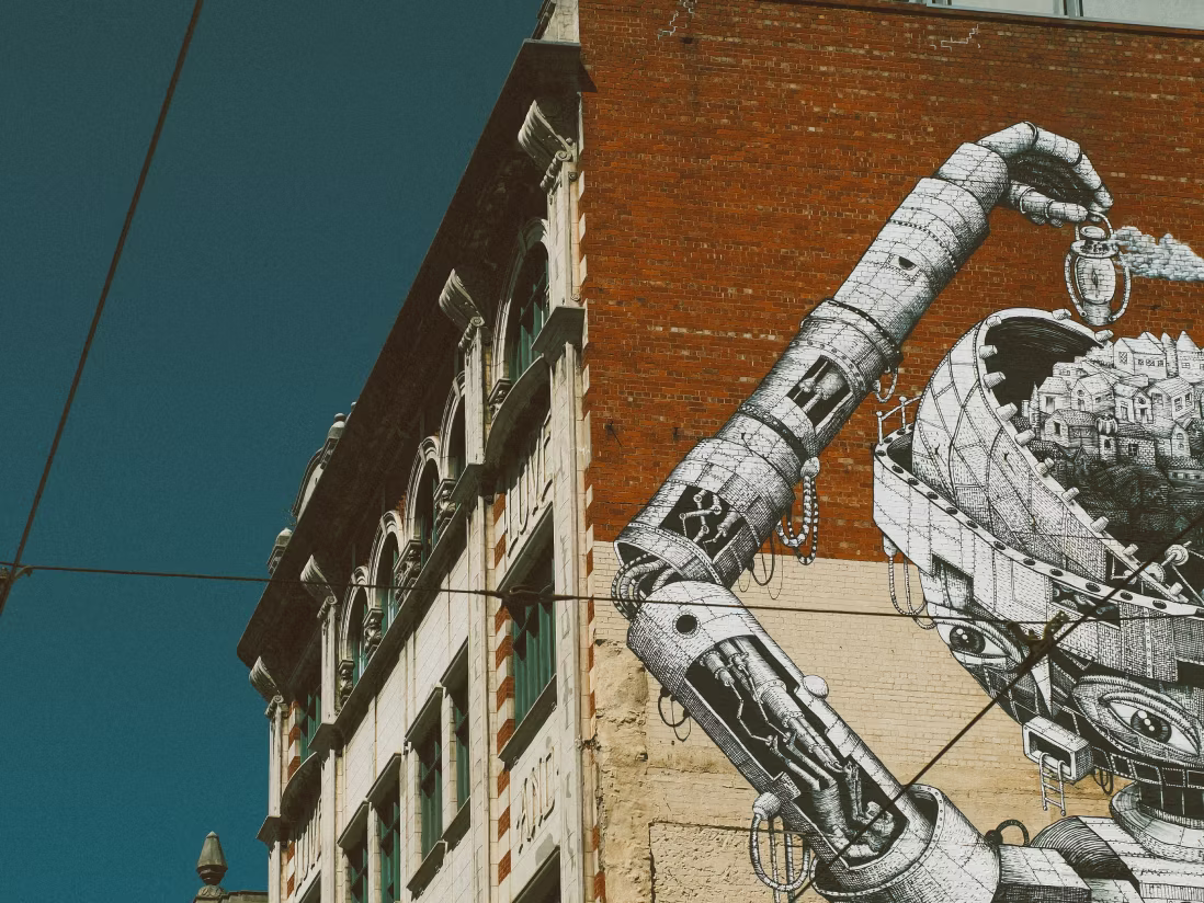 Mural on a brick building wall featuring a detailed robotic arm holding a pocket watch above a mechanical head filled with small houses.