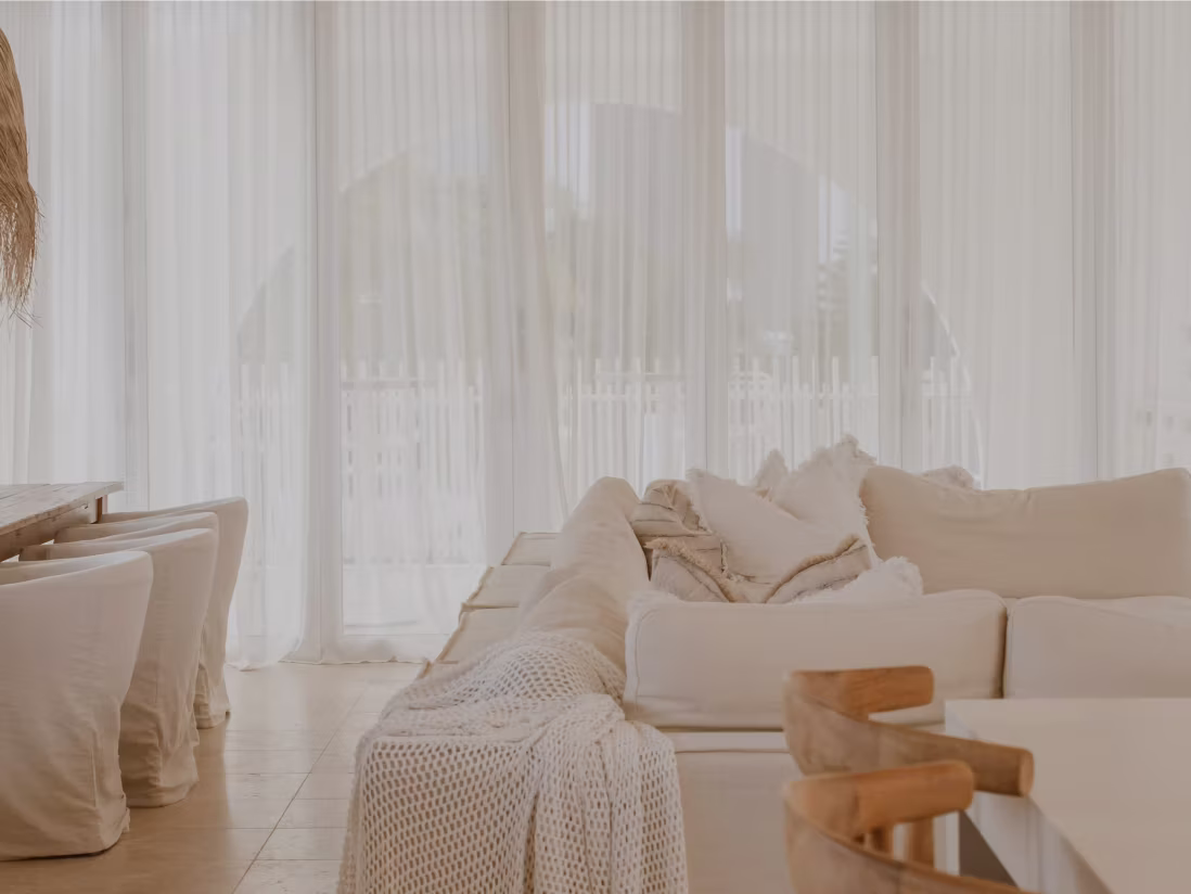 Bright living room with large beige sectional sofa, soft pillows, and sheer white curtains over tall windows.