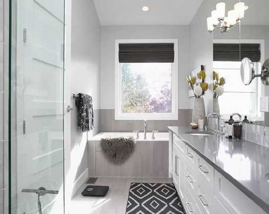 Modern bathroom with white walls, a built-in bathtub under a window, gray countertop vanity with double sinks, patterned black and white rug, and a glass shower door.