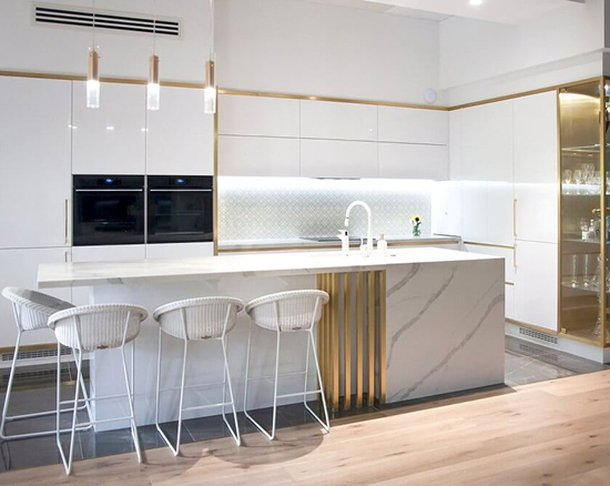 Modern white kitchen with marble island, four white bar stools, built-in ovens, and gold-accented cabinetry.