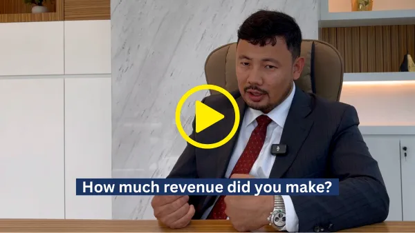 Man in a suit sitting at a desk with caption 'How much revenue did you make?' and a yellow play button overlay.