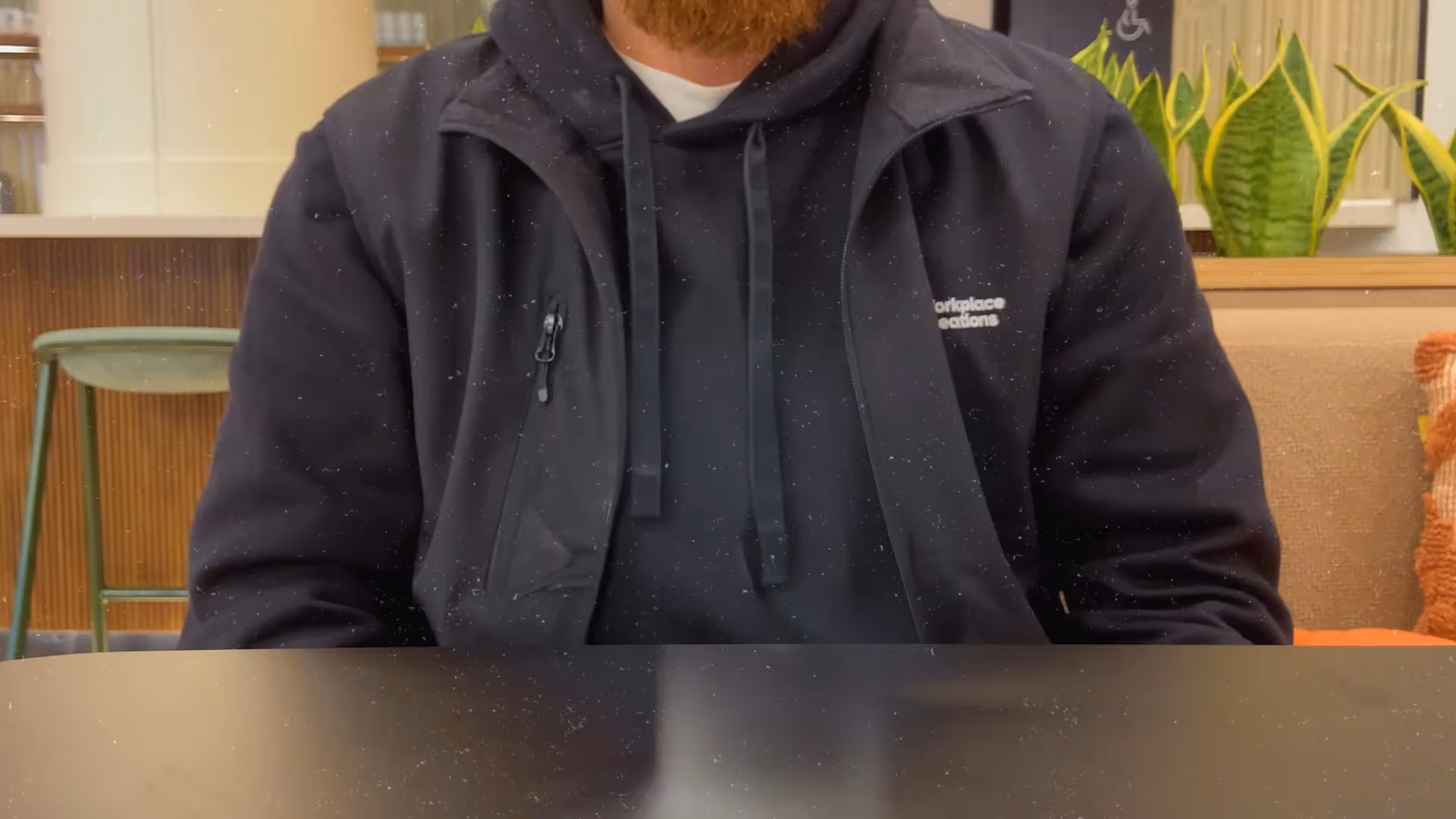 Person wearing a black hoodie and jacket sitting at a table in a cozy indoor space with plants.