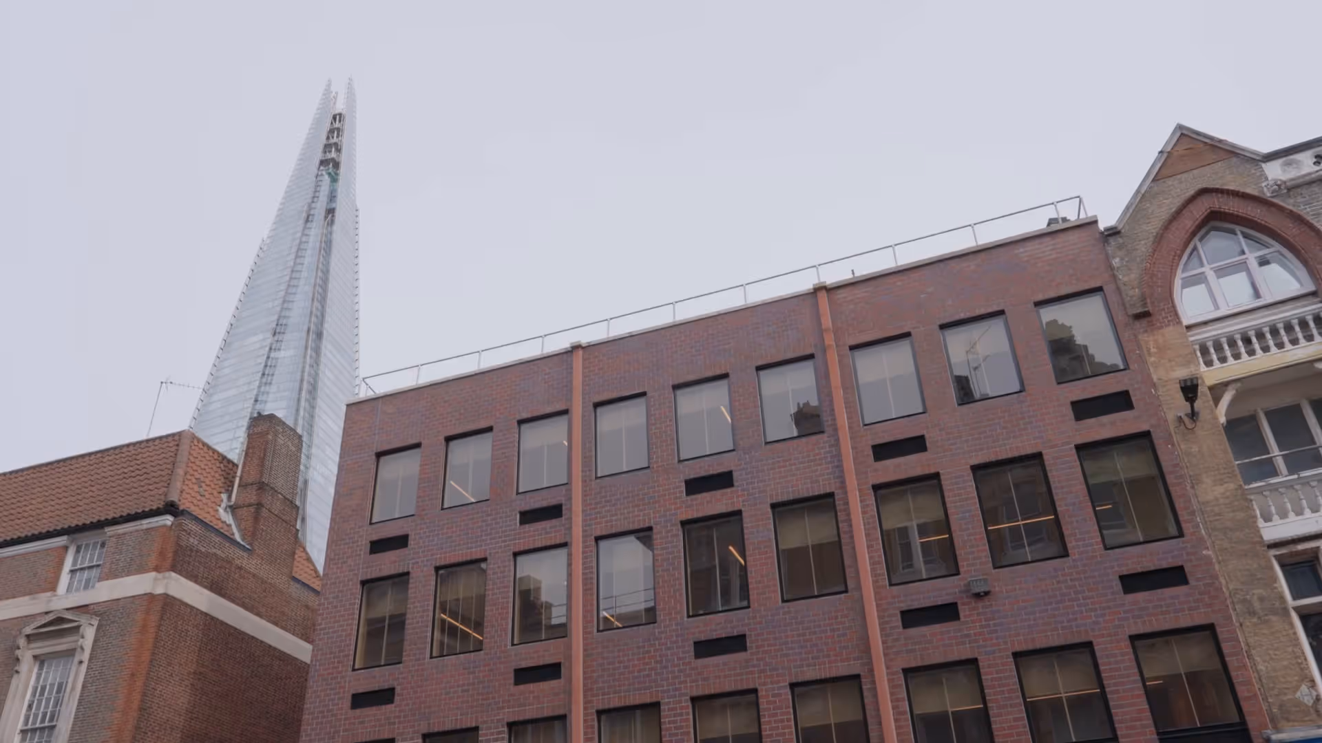 Modern tall glass skyscraper behind older brick and stone buildings under a gray sky.