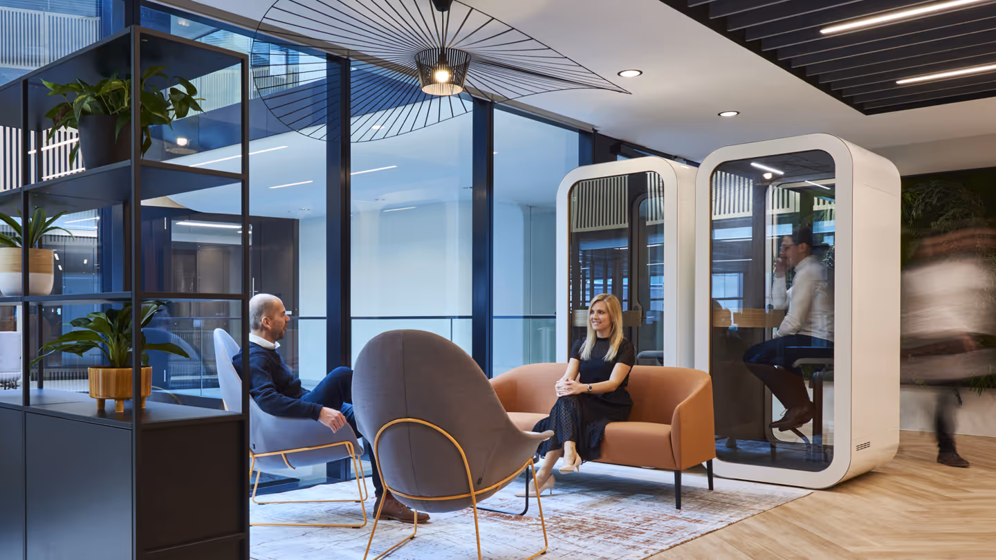 Modern office lounge with two people seated and private pods with workers inside beside large windows.