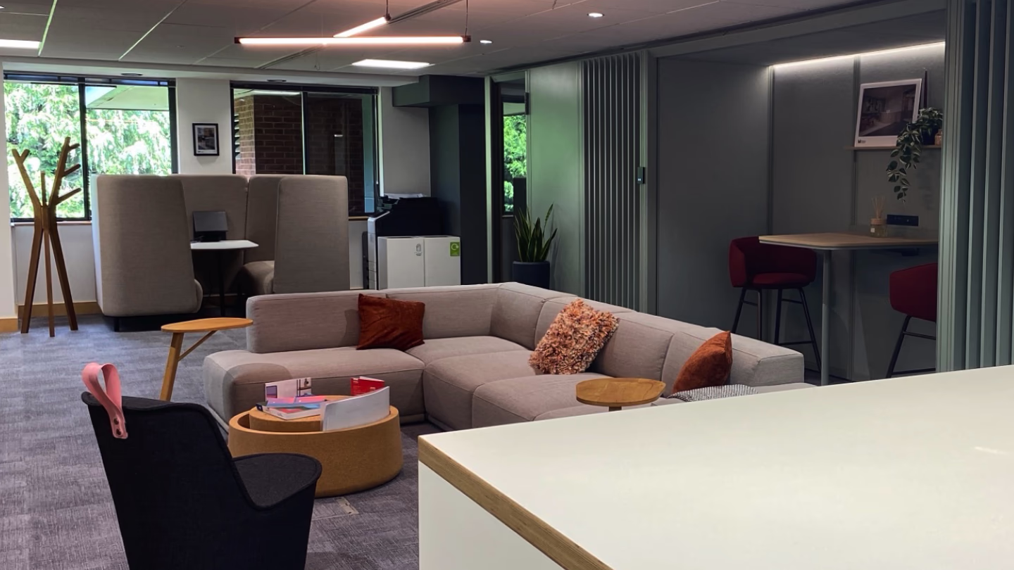 Modern office lounge with beige sofa, orange pillows, round wooden tables, and partitioned seating areas.