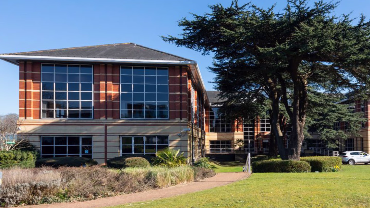 Large brick building with wide windows, surrounded by grass, shrubs, and a tall tree on a sunny day.