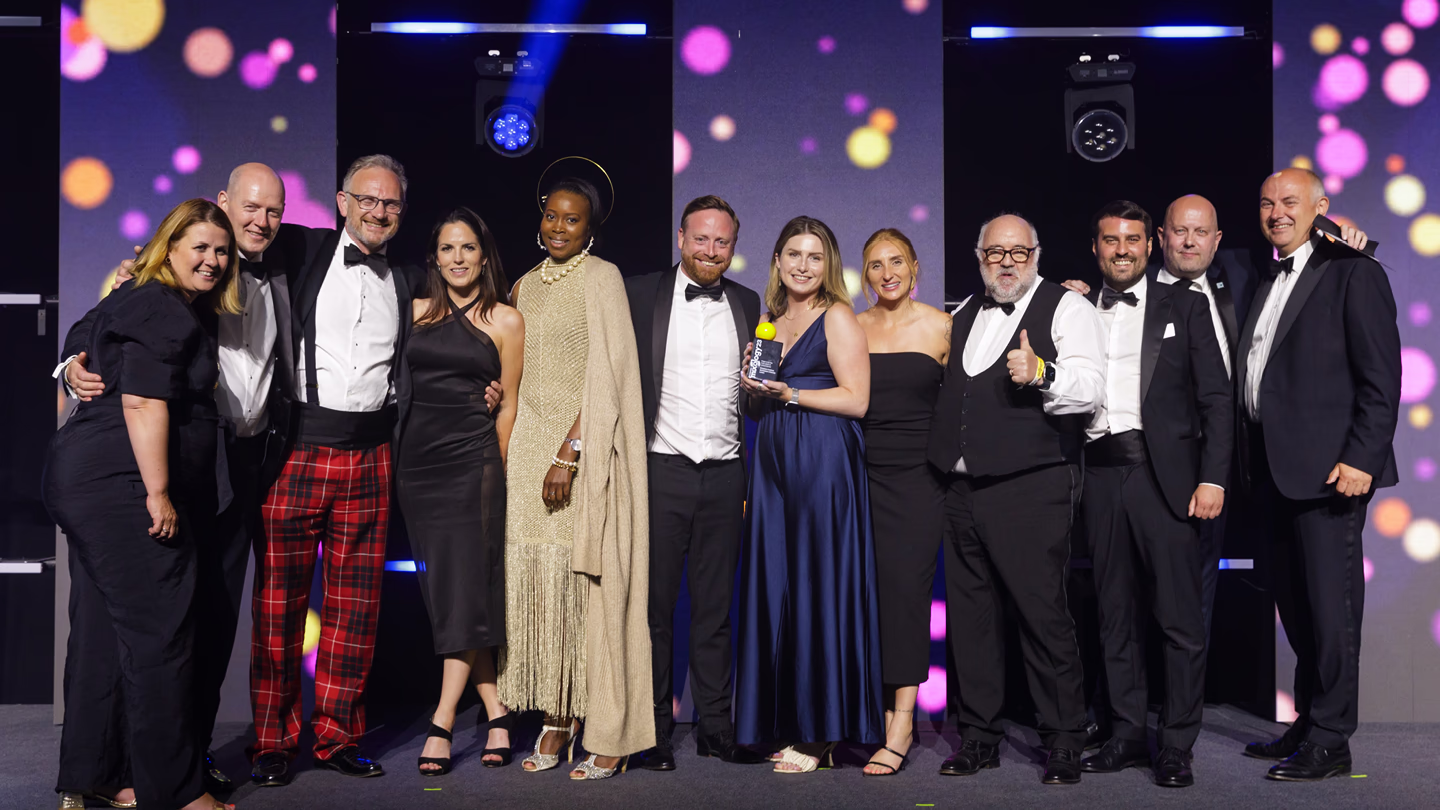 Group of formally dressed people posing on stage with an award, colorful lights in background.