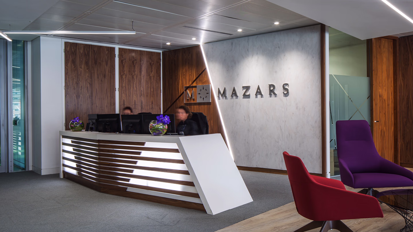 Modern office reception with illuminated desk, two employees, and Mazars logo on wall.