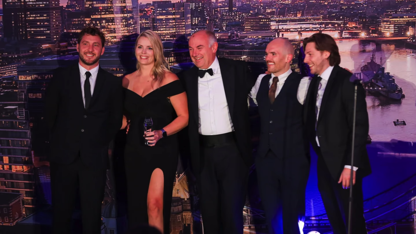 Five people dressed in formal attire smiling and posing for a photo against a cityscape backdrop.