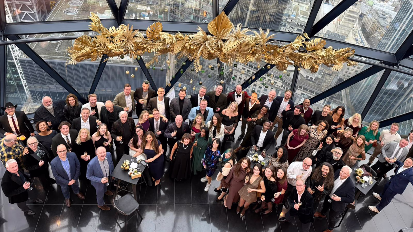 Large group of diverse adults posing indoors under golden decorations with city views through glass walls.