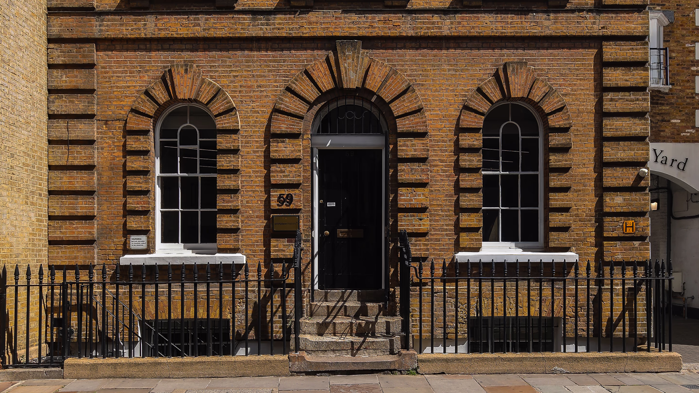 Brick building facade with black door, two arched windows, and black metal railing.