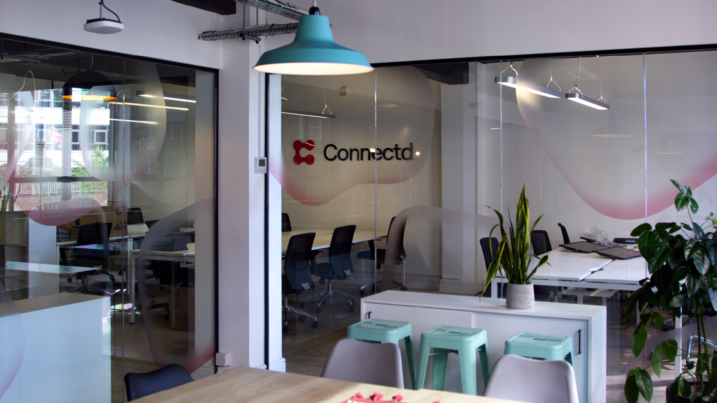 Modern office space with glass walls, conference room, green stools, plants, and pendant light.