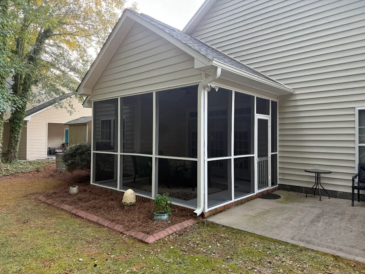Backyard view of a beige house with a screened-in porch and a small concrete patio with a chair and round table.