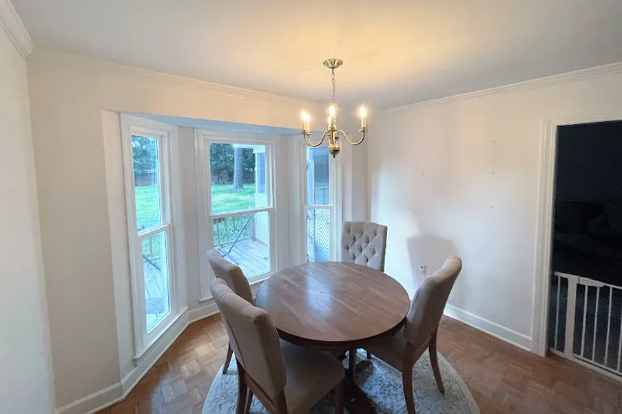 Cozy dining area with a round wooden table, four cushioned chairs, a chandelier, and large bay windows overlooking a green yard.