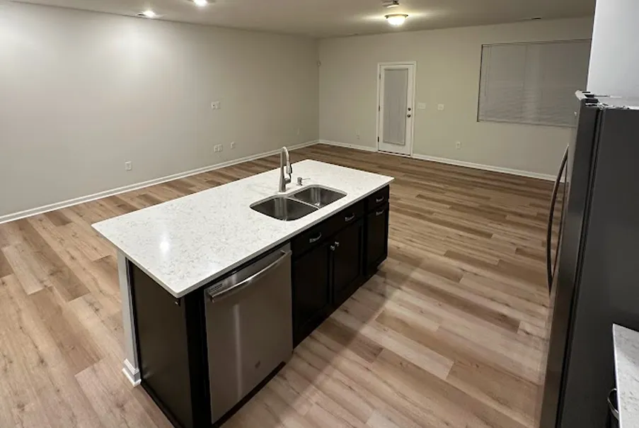 Modern kitchen with a large island featuring a white countertop, stainless steel sink and dishwasher, and brown wood flooring.