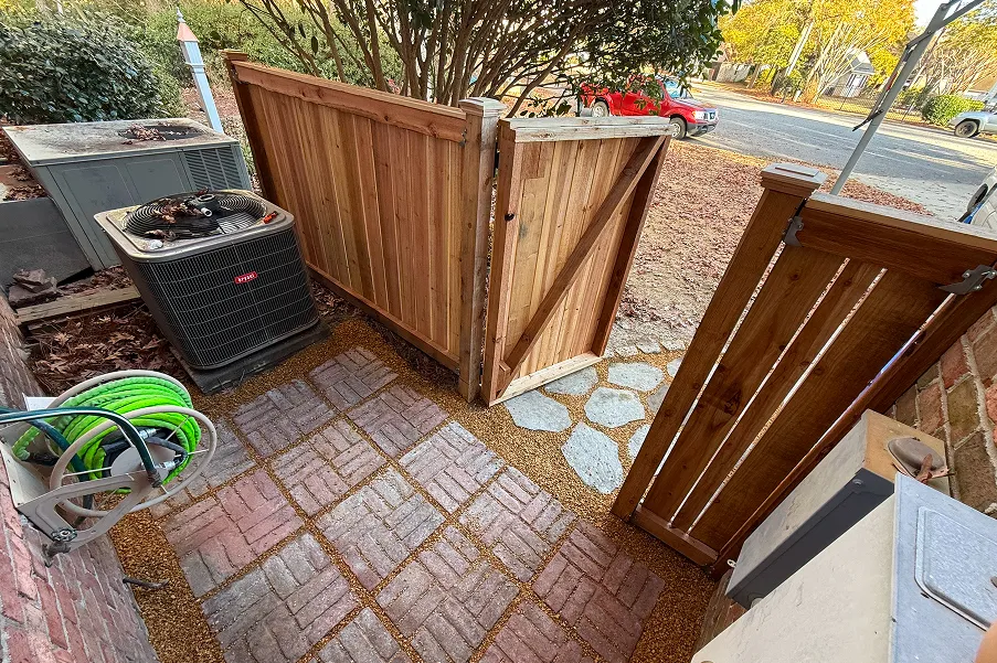 Wooden gate open beside brick wall and air conditioning unit with garden hose coiled on a reel.