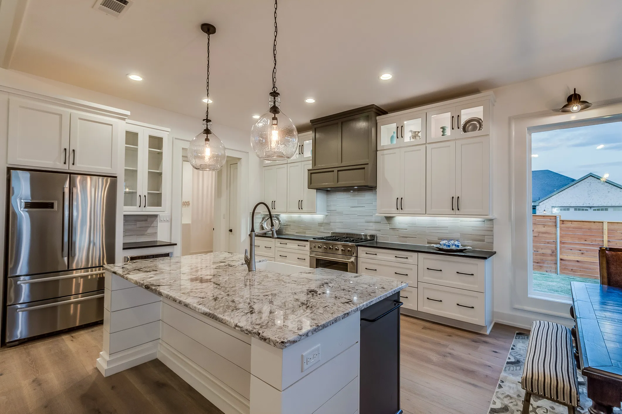 Modern kitchen with white cabinets, marble island countertop, stainless steel refrigerator, and glass pendant lights.