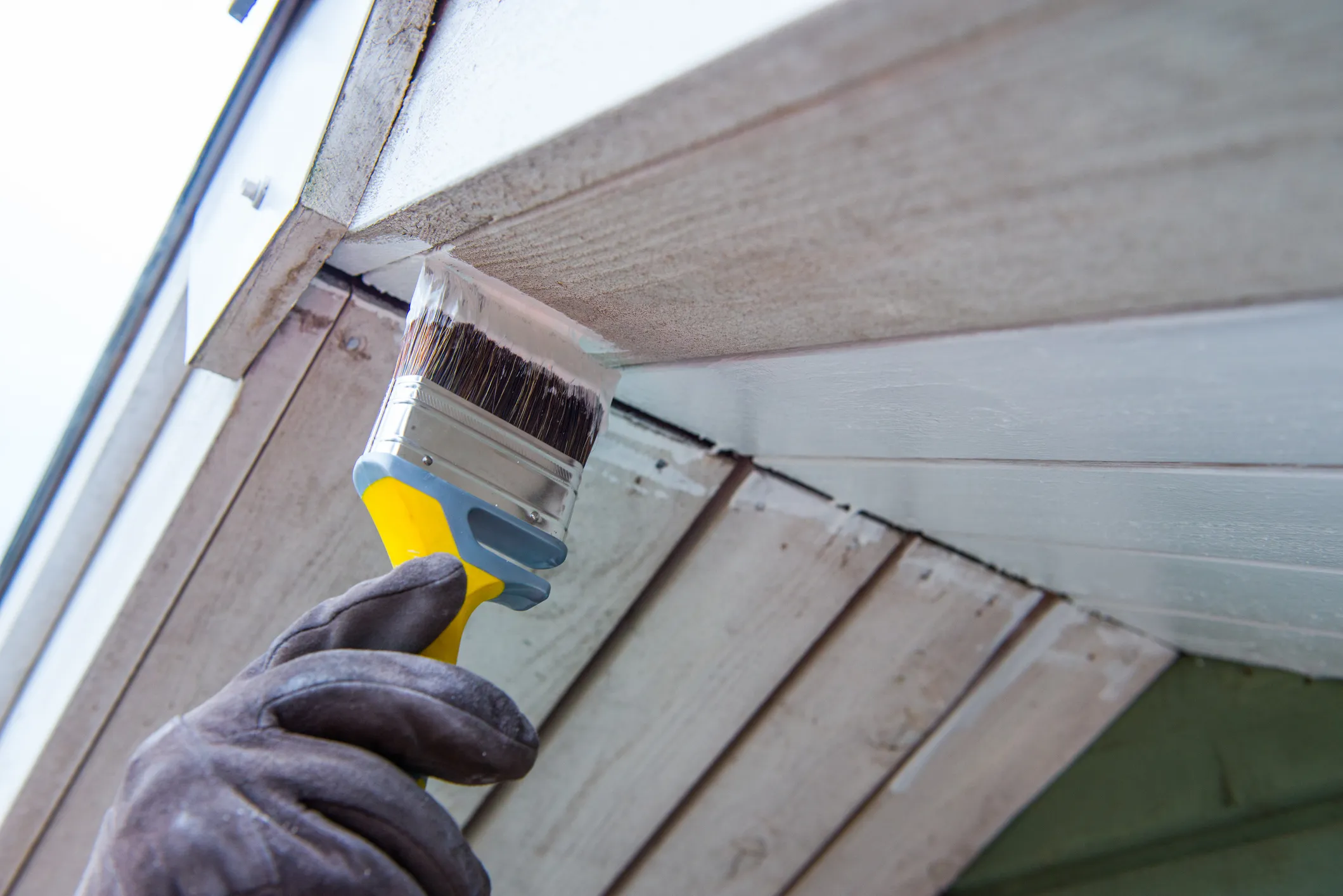 Hand in glove holding a yellow paintbrush painting white trim on an exterior wooden surface.