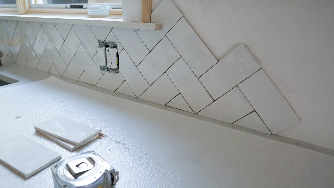 Kitchen backsplash installation in progress with white herringbone tiles and loose tiles on countertop near a tape measure.