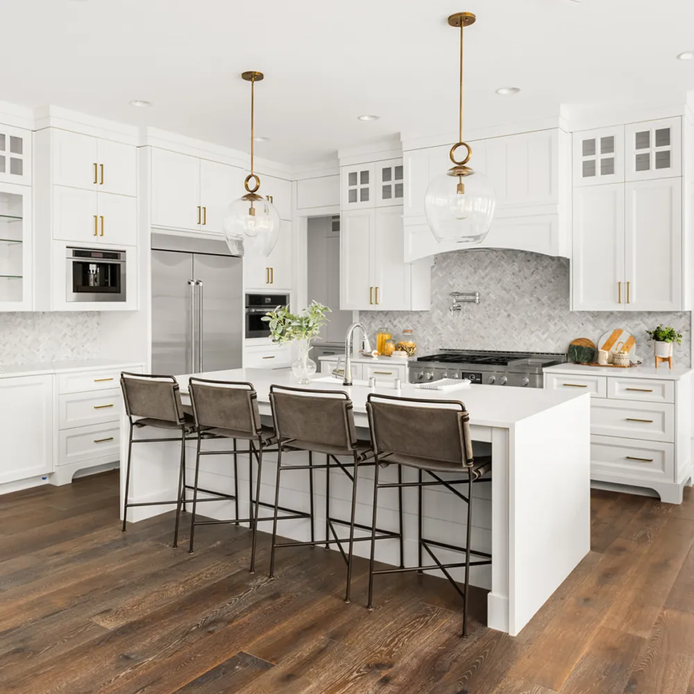 Modern white kitchen with large island, four brown bar stools, pendant lights, and stainless steel appliances.
