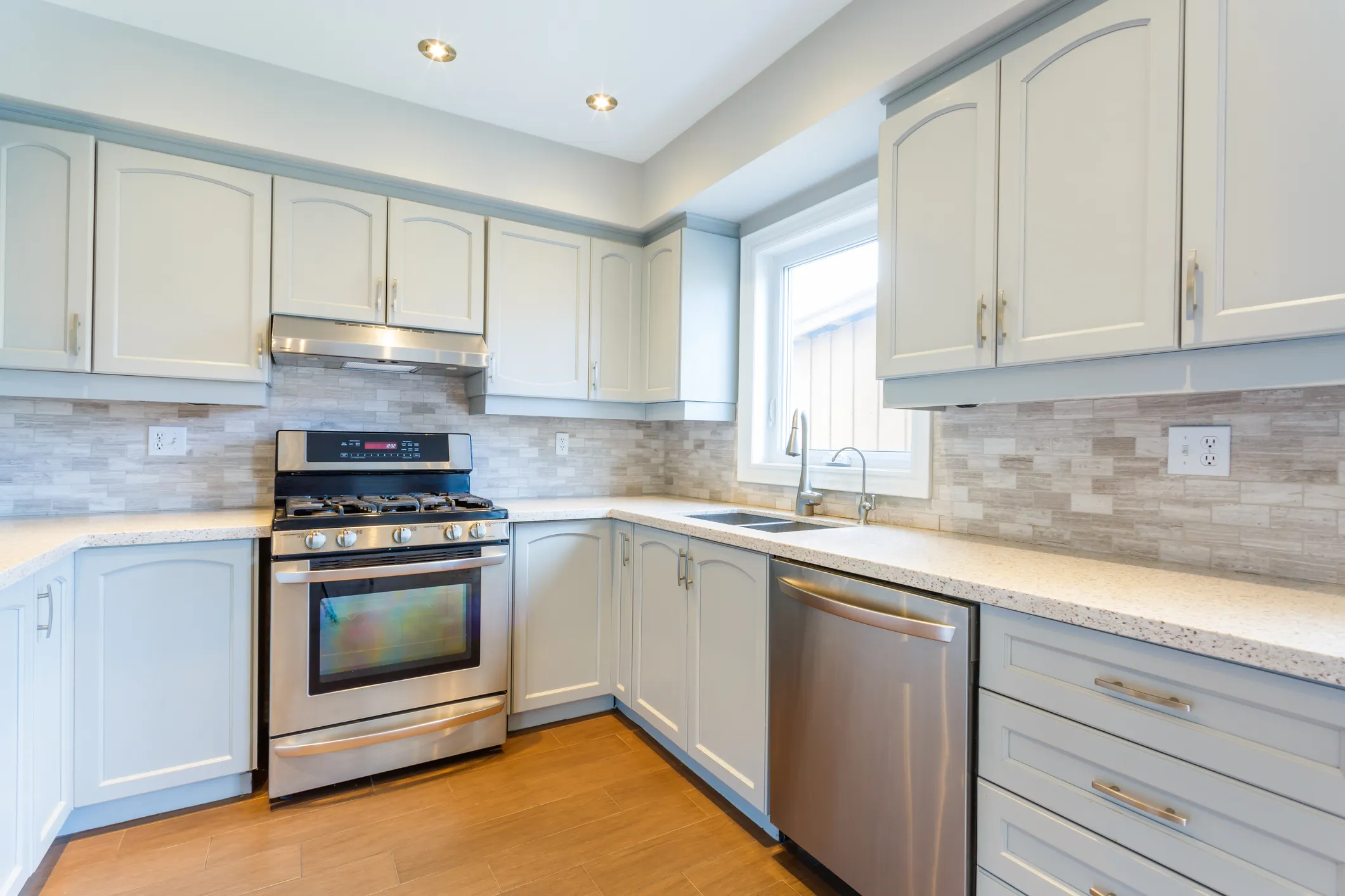 Bright modern kitchen with white cabinets, stainless steel gas stove, dishwasher, and a window above the double sink.