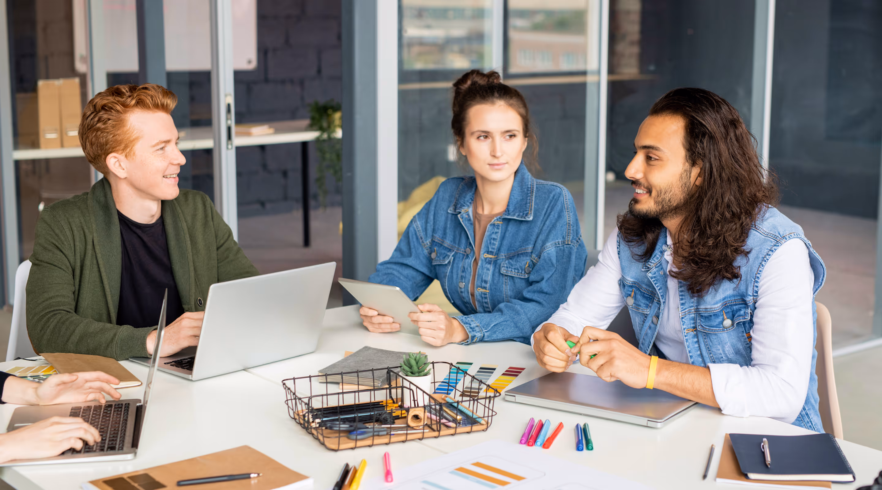 Three young adults in a meeting room collaborating with laptops, a tablet, and colorful markers on a white table.