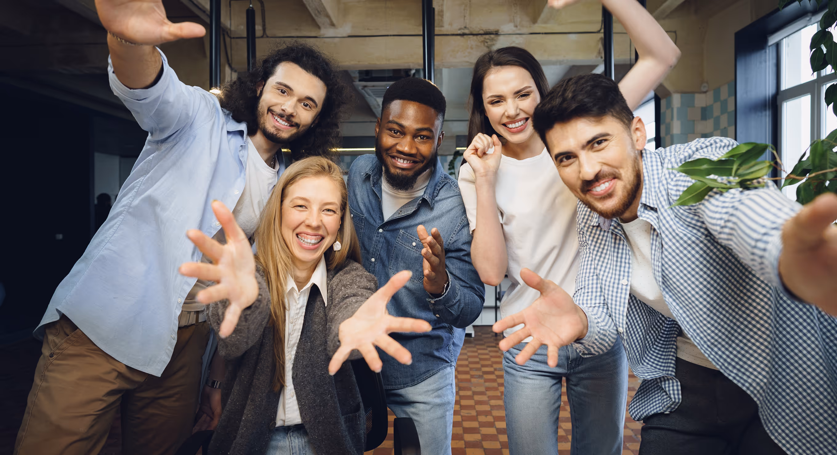 Five diverse young adults smiling and reaching their hands toward the camera in a bright indoor space.