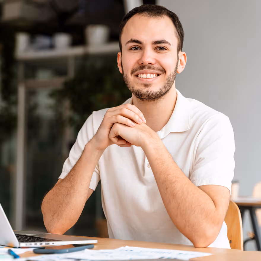 Young man in a white polo shirt smiling with hands clasped sitting at a desk with a laptop and documents.
