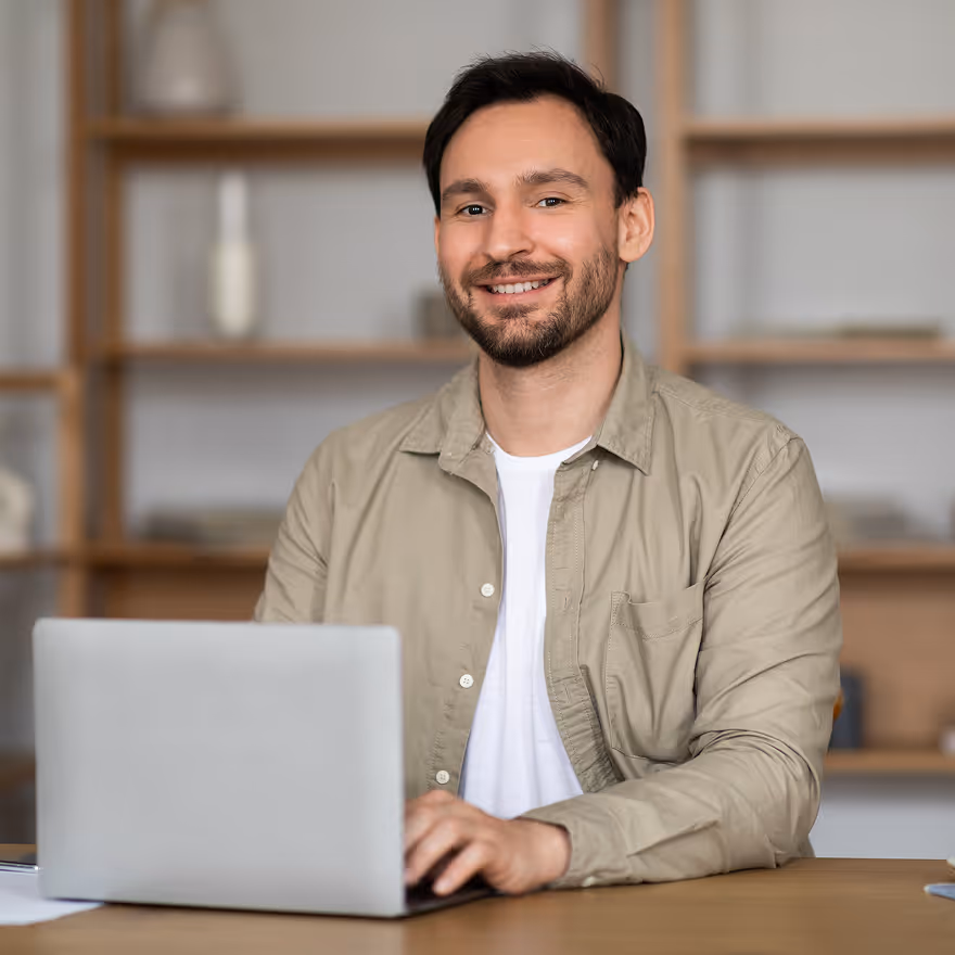 Smiling man with dark hair and beard sitting at a desk and using a laptop in a modern room.