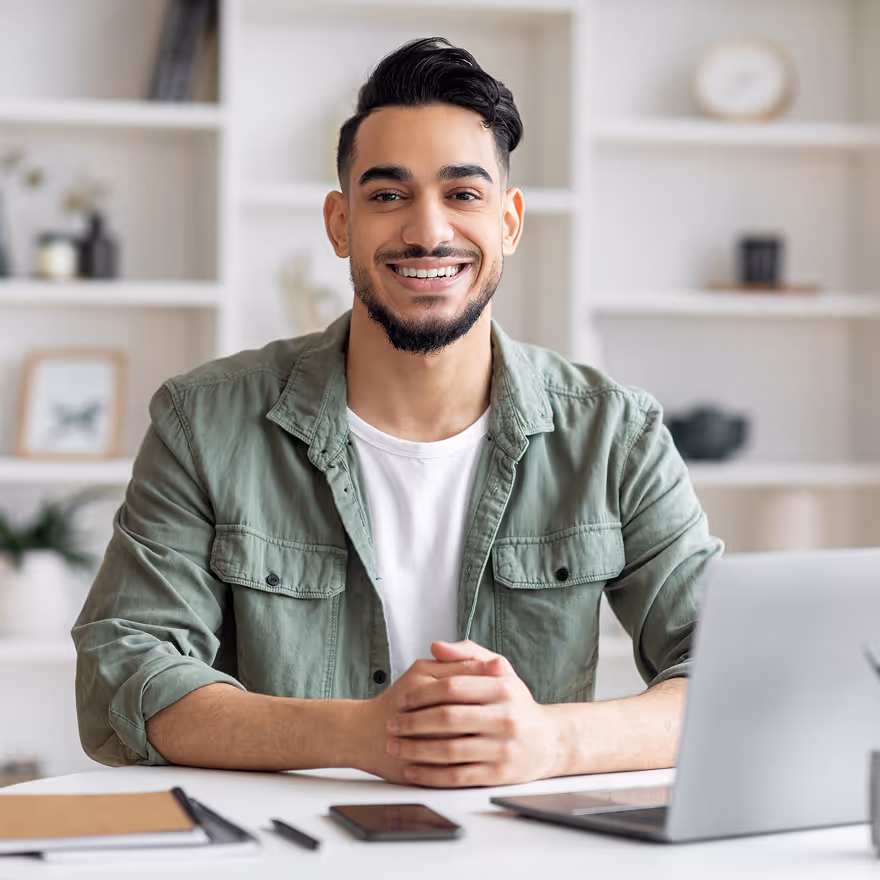Smiling young man with dark hair and beard sitting at a desk with a laptop, phone, and notebook in a bright room with shelves in the background.