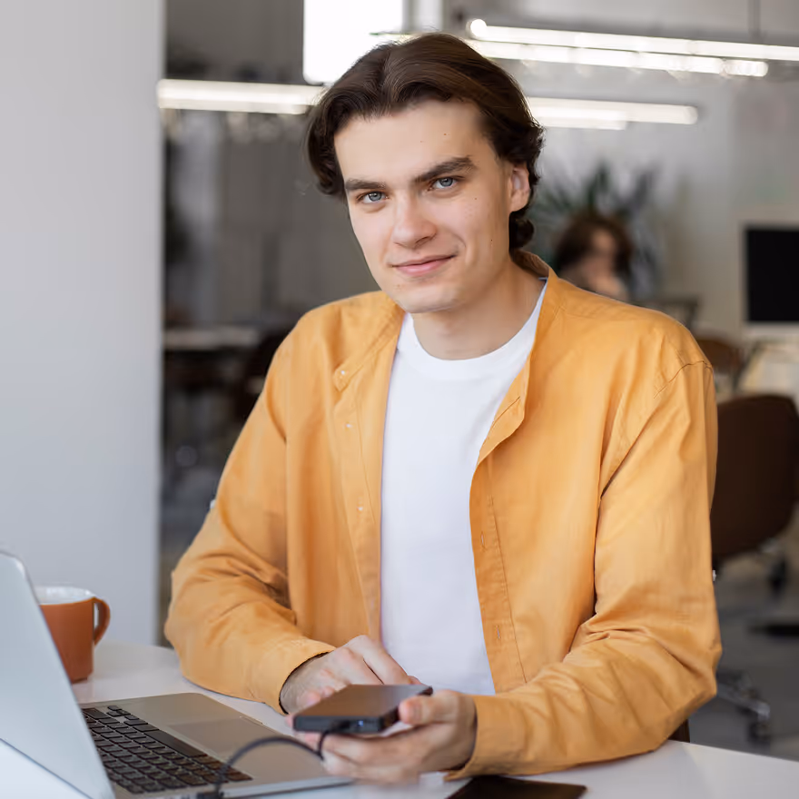 Young man in a yellow shirt sitting at a desk with a laptop, holding a smartphone and smiling.