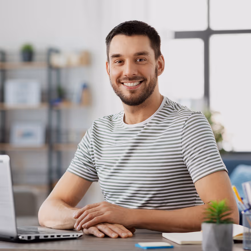 Smiling man with short dark hair and beard wearing a striped t-shirt sitting at a desk with a laptop and notebook.
